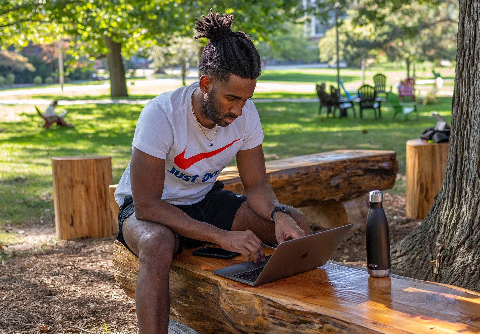 A student on their laptop sits on a wooden bench under a tree on a sunny day